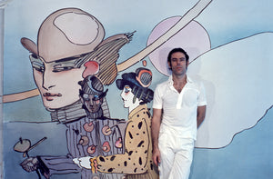 Peter in white pants and white shirt leaning against a gigantic painting of woman that he created.