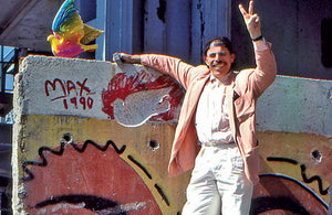 Peter Max standing on a ladder near the top of a piece of the Berlin Wall. To his right is his signature painted on the wall and a statue of a dove that he carved from it.