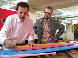 Peter Max and Ringo Starr standing over a painted piano while Peter signs it with a paintbrush. 